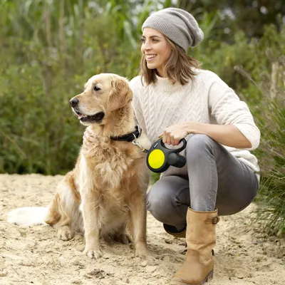 Nő egy sárga Flexi-Rollleine pórázt tart, amely egy ülő Golden Retrieverhez van rögzítve.
