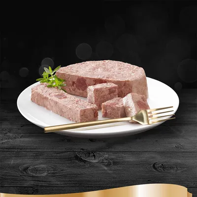 Куски мясного паштета на белой тарелке с золотистой вилкой и веточкой зелени, черный фон. Вид продукта без упаковки.