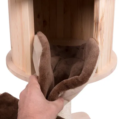 Hand plaatst een bruin kussen in een ronde houten kattenhuis. Kussen van stof, huis van licht hout zichtbaar.