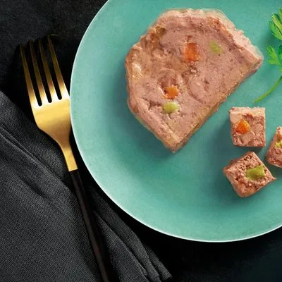 Plak natvoer met zichtbare stukjes wortel en erwt op een turquoise bord, enkele blokjes afgesneden. Gouden vork en zwarte servet naast het bord.