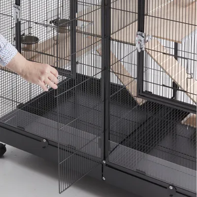 Grosser Metallkäfig mit Gittertüren, Holzplattformen und Futterbehältern. Hand öffnet eine der Türen. Käfig auf Rollen für einfache Bewegung.