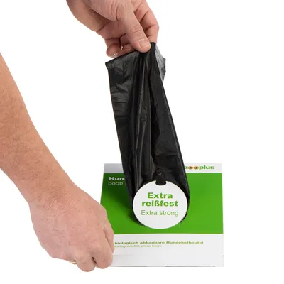 Hand pakt een zwarte hondenpoepzak uit een groene en witte doos met tekst: Extra reißfest, Extra strong, biologisch afbreekbare Hundekotbeutel, zooplus.