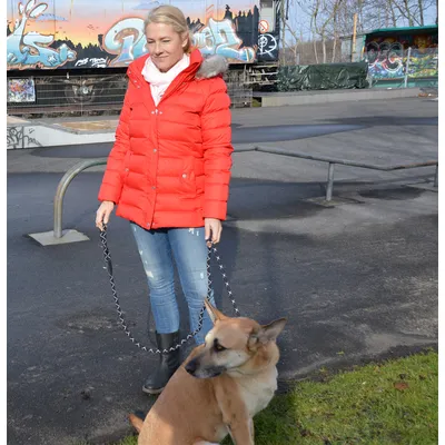 Woman in a red winter jacket holding a dog on a black and white patterned lead on a pavement with graffiti in the background.