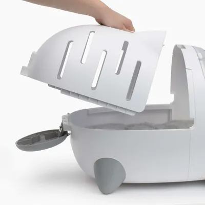 En hånd løfter låget af en hvid kattetoilet med aftagelig overdel og sideklap, indersiden med synlig strøelse.