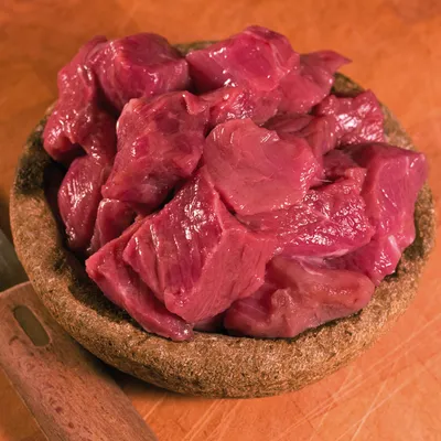 Bucăți de carne crudă roșie tăiate cuburi, așezate într-un bol maro pe o suprafață din lemn. Nu este vizibil niciun ambalaj sau marcă.