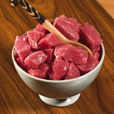 Bucăți de carne crudă roz în bol ceramic, amestecate cu lingură din lemn. Nu este vizibil niciun brand sau etichetă pe produs.