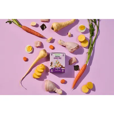 Box of grain free puppy food surrounded by visible ingredients including chicken drumsticks, carrots, parsnips, sweet potato, hard-boiled egg, and vegetable pieces.