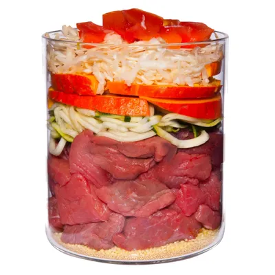 Glass container with visible layers of raw beef chunks, spiralised courgette, sliced carrot, shredded cabbage, and diced tomato on top of yellow grains. No brand or text visible.