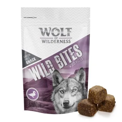 Wolf of Wilderness Wild Bites Wild Hills, snack senza cereali. Confezione con immagine di lupo e cubetti marroni visibili accanto alla busta. Simbolo anatra in basso a sinistra.
