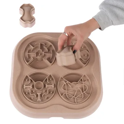 Plateau interactif en plastique beige avec quatre labyrinthes ronds, main soulevant une pièce en forme d’os. Accessoire pour animaux visible sans marque ni texte.