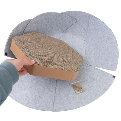 Mano sostiene un rascador marrón de cartón rodeado de fieltro gris. Bolsa con hierba gatera al lado.