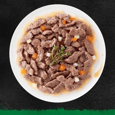 Ciotola bianca con bocconcini di carne in salsa, pezzi di carota e patata visibili, decorazione con rametto di erbe aromatiche al centro.