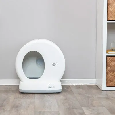Wit, rond kattenbakapparaat met digitaal bedieningspaneel en zichtbaar netsnoer, geplaatst op een houten vloer tegen een grijze muur. Merknaam 'PETKIT' zichtbaar op de voorkant.