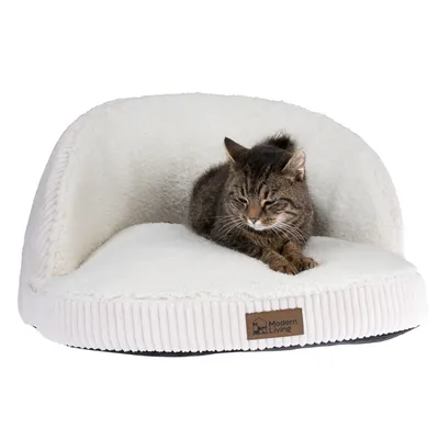 Katzenbett s bílým plyšovým potahem a hnědým štítkem 'Modern Living'. Kočka na něm leží uvolněně.