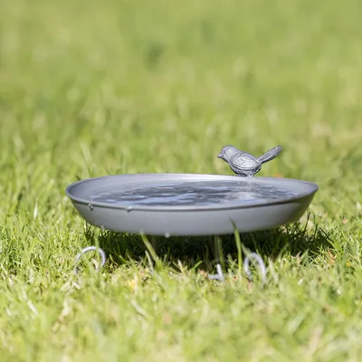 Grau lackierte Vogeltränke aus Metall mit dekorativer Vogelfigur, auf einem Rasen platziert.