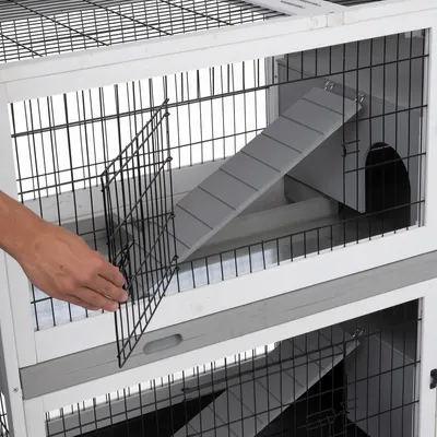 Cage à petits animaux en métal et bois blanc, porte grillagée ouverte, rampe grise menant à un abri intérieur. Main ouvrant la porte. Deux niveaux visibles.