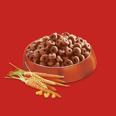 Червена купа за храна с кафяви рингови гранули, до нея моркови, пшеница и царевични зърна на червен фон.