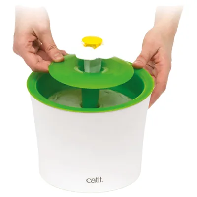 Persoon toont het monteren van de groene deksel op een witte drinkfontein met gele bloemopzet van het merk Catit.