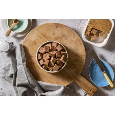 Bowl filled with cubed pieces of pet food on a wooden board, with additional cubes and a block of the same food shown beside it.