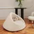 Cat bed made of white fabric with enclosed cave shape, cat sleeping inside. Environment: wooden floor, black table with vase and plants.