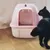Затворена котешка тоалетна в розово с прозрачна люка и лого Trixie, до нея черна котка.