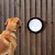 Perro mirando una ventana redonda en una valla de madera. El producto muestra una opción de instalación de ventana para mascotas.