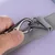 Mousqueton en métal argenté attaché à une boucle en tissu gris, maintenu par une main, sur fond violet. Détail du système d’attache pour laisse ou harnais.