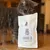 Chat gris à côté d’un sachet blanc marqué « Kater Kasimir Catnip » avec illustration de chat et photo d’une femme sur l’étiquette. Herbe à chat visible au sol devant le sachet.