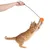 Varita de juego con bola de peluche naranja, sostenida por una mano. Gato salta y atrapa la bola.