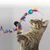 Chat tigré jouant avec une baguette à plume et boule bleue, ornée de spirales colorées en tissu. Jouet visible en main du chat, matériaux : tissu, plastique, plumes.