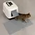 Kattenbak met witte kap, transparante klep met pootafdrukken en grijze mat ervoor; kat loopt uit de kattenbak over de mat.
