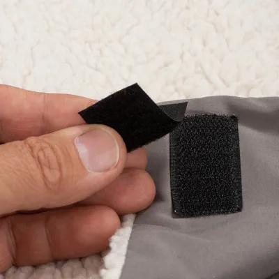 Detail van een hand die een zwarte klittenbandsluiting opent op grijze stof met witte fleecevoering, bevestiging zichtbaar.