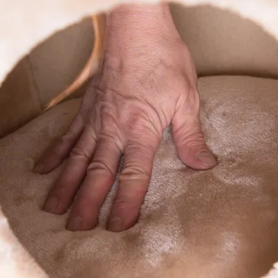 Hånd trykker på blød, beige pude i plysmateriale inde i et kæledyrshus. Materialets struktur og polstring vises tydeligt.