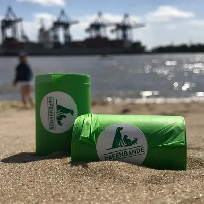 Зелени ролки за кучешки изпражнения с лого Hafenbande, поставени в пясъка на плажа. На заден план пристанищни кранове и вода.