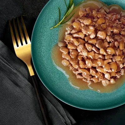Assiette verte contenant des morceaux de nourriture humide en sauce, garnie d'une branche de romarin. Fourchette dorée posée à côté sur une serviette noire.