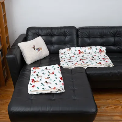 Manta branca para cão com motivos coloridos de cães e design natalício, estendida num sofá de couro preto. Almofada a condizer com bordado de cão ao lado.