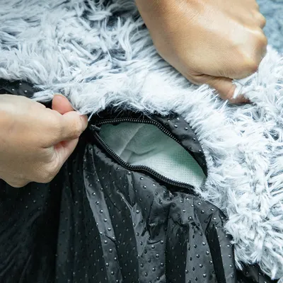 Nahaufnahme eines Reissverschlusses an einem flauschigen, grauen Stoffbezug mit schwarzer Unterseite. Zeigt die Möglichkeit, den Bezug zu öffnen und zu entfernen.