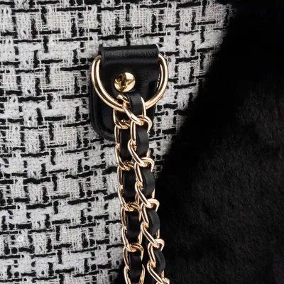 Detail van een goudkleurige metalen ketting met zwart kunstleer, bevestigd aan een ring op een tas van zwart-wit geweven stof en zwarte pluche stof.