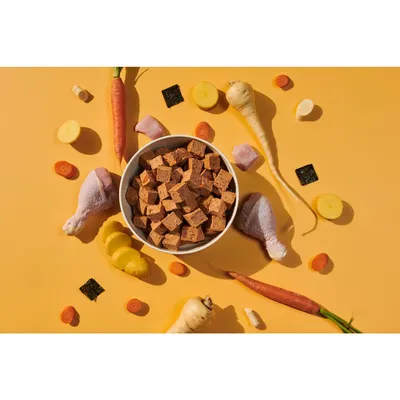Bowl of cubed pet food surrounded by raw chicken drumsticks, carrot, parsnip, potato slices, and small squares of seaweed, showing visible ingredients used in the product.