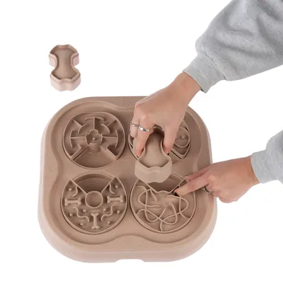 Interaktivt brädspel för husdjur i beige plast med fyra olika labyrintmönster och benformade delar, två händer visar hur delarna flyttas på spelplanen.
