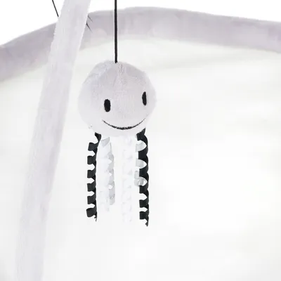 Jouet suspendu en peluche grise en forme de méduse avec visage souriant, tentacules en rubans noirs et blancs, accroché à une arche recouverte de tissu.