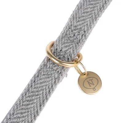 Graues Halsband aus Stoff mit goldfarbenem Metallring und Anhänger mit eingraviertem 'NT'-Logo.