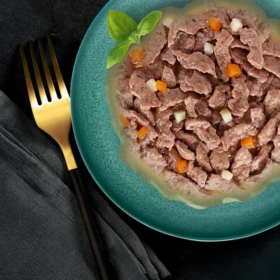 Chunks of wet pet food with visible orange and white vegetable pieces in gravy, served on a green plate with a basil garnish. Gold fork and dark napkin beside the plate.