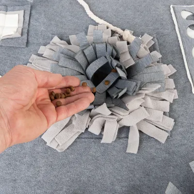 Main versant des croquettes dans un tapis de fouille en tissu gris et beige, avec lanières épaisses et corde visible sur le côté.