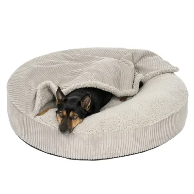 Rond, beige huisdierenbed met zachte rand en deken, het dier ligt half onder de deken in het bed.