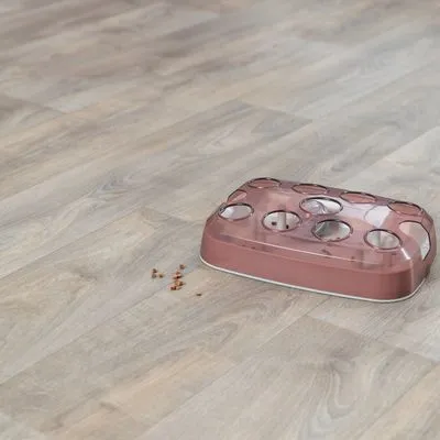 Interactief voerspeelgoed van roze plastic met meerdere ronde openingen, enkele brokjes voer liggen ernaast op de vloer.
