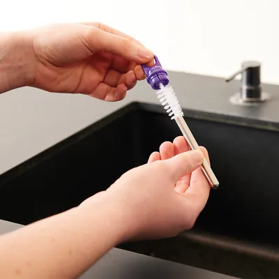 Kéz egy fogantyús, sörtés tisztítókefe használatát mutatja egy fém szívószál tisztításához mosdókagyló felett.