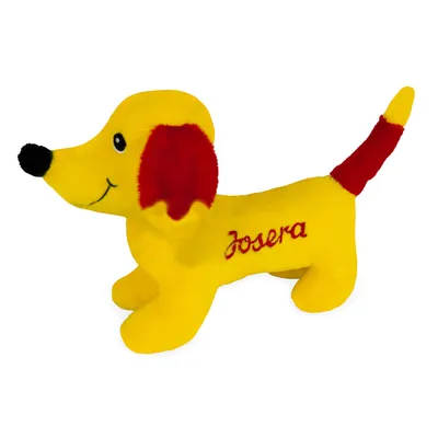 Yellow plush toy in the shape of a dog with red tail and ears, embroidered with the inscription 'Josera'.
