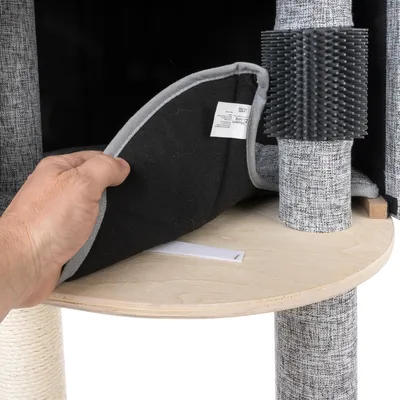 Main soulevant un coussin amovible en tissu noir avec étiquette sur une plateforme en bois d’un arbre à chat, poteaux recouverts de tissu gris et brosse noire fixée à un poteau.