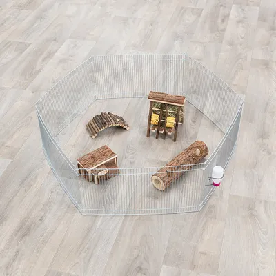 Achtseitiges Metallgehege mit Holzspielzeug, Heuraufe und Trinkflasche auf hellem Laminatboden.